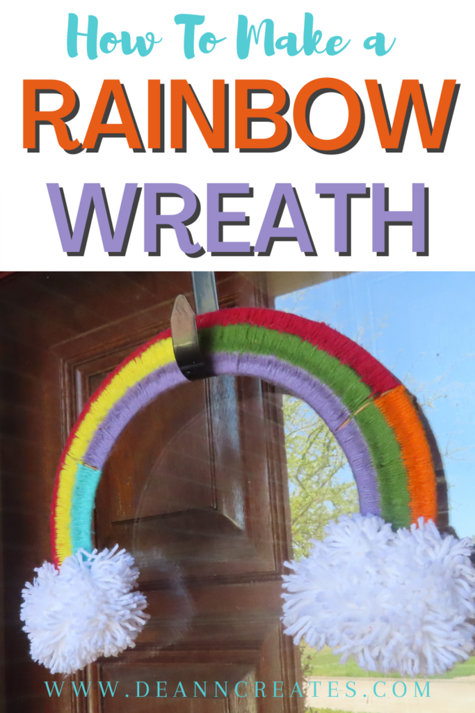 This image shows a Pinterest pin entitled "How to Make a Rainbow Wreath." The picture shows a yarn woven rainbow wreath with white pom poms for clouds hanging in front of a wooden door.