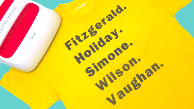 This picture shows a yellow shirt with the names of jazz singers on it and a Cricut EasyPress 2 (white with red accents) sitting on the shoulder of the shirt. The background is turquoise