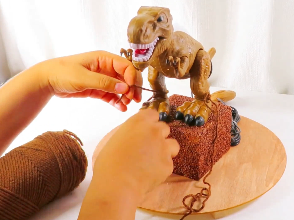 Tying brown yarn around the dinosaur's ankles to secure it to the centerpiece