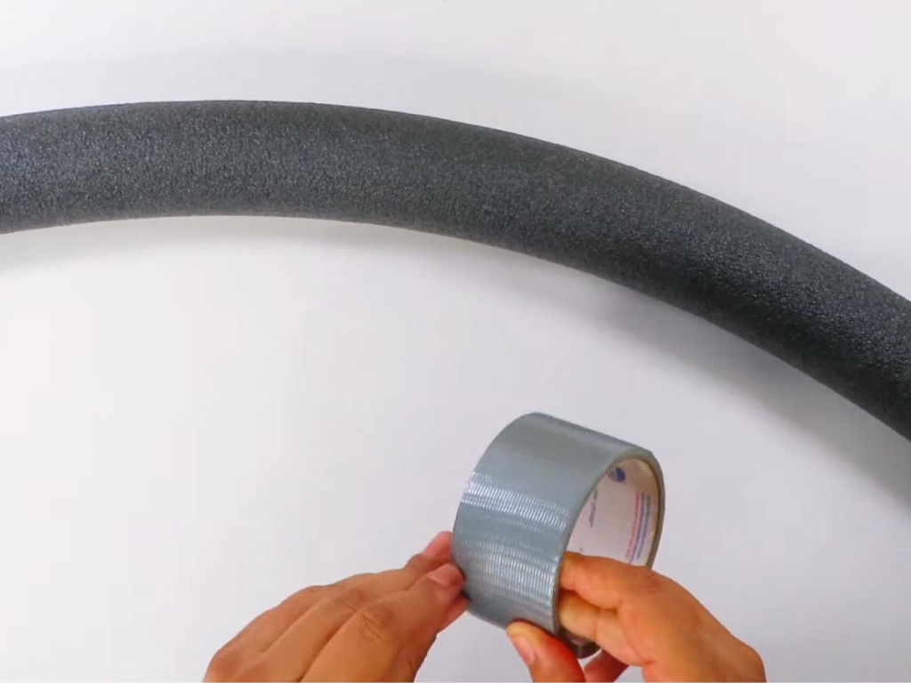 This image shows a pair of hands holding silver duct tape with a black pool noodle laying on the surface behind it.
