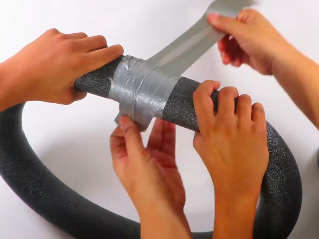 This image shows a pair of hands holding the ends of a pool noodle together while another pair of hands wraps silver duct tape where the pool noodle ends meet.