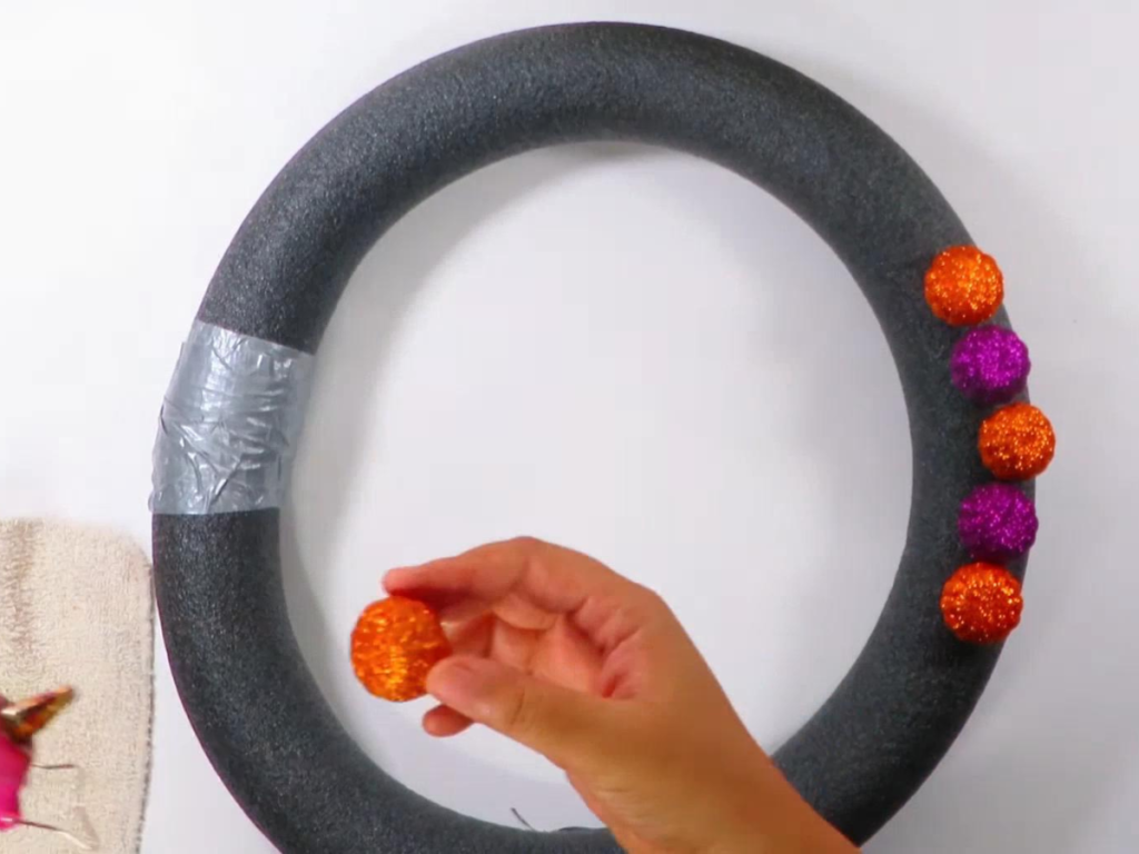 This image shows a pool noodle wreath held together by silver duct tape with five purple and orange pumpkin fillers are glued to the wreath.