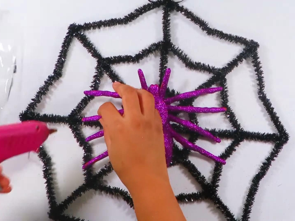 This image shows a hand lifting up the purple glitter spider's legs to place a dollop of hot glue underneath..