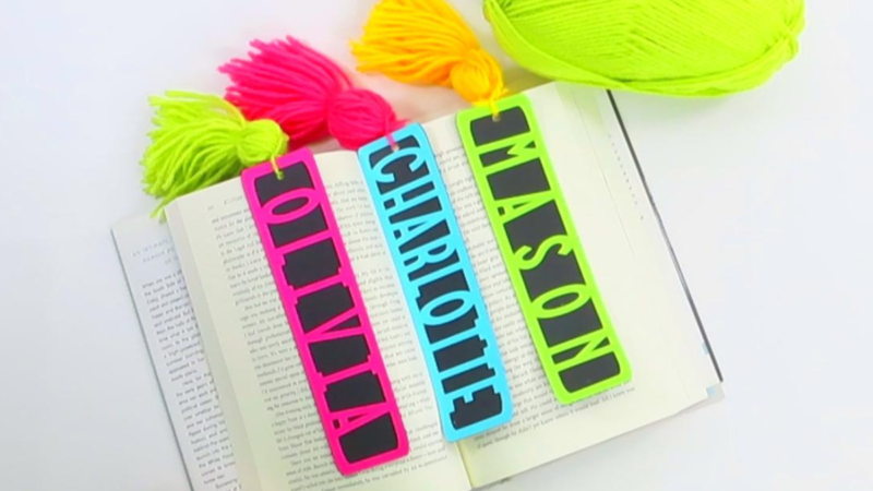 This image shows an open book with three colorful name DIY Bookmarks with yarn tassels laying across the pages