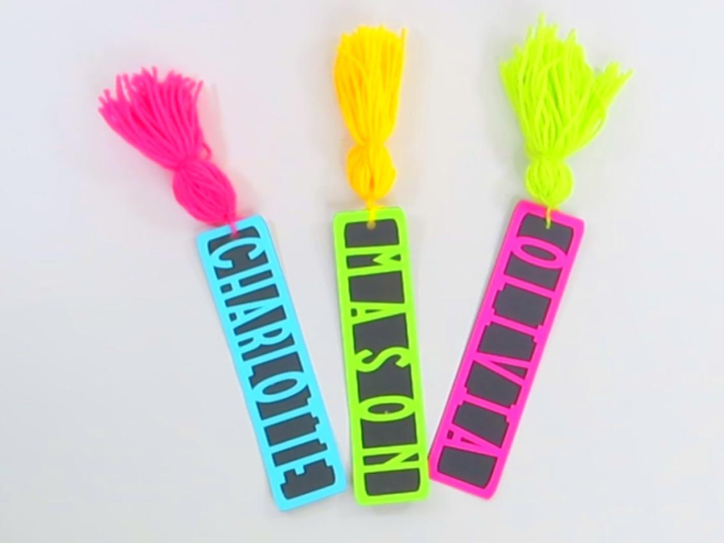 This image shows the colorful DIY Bookmarks laying side by side. All three of them have a black base. From left to right the Charlotte bookmark has a blue top layer with a pink tassel. The Mason bookmark has a green top layer with a yellow tassel and the Olivia bookmark has a red top layer with a green tassel.