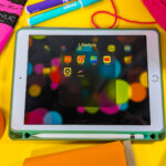 This image shows a white iPad inside of a turquoise case surrounded by colorful craft supplies including orange duct tape, orange fabric, green felt, pink yarn, pink and black tubes of paint and turquoise and purple paint markers