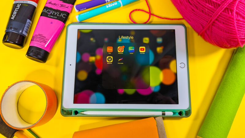 This image shows a white iPad inside of a turquoise case surrounded by colorful craft supplies including orange duct tape, orange fabric, green felt, pink yarn, pink and black tubes of paint and turquoise and purple paint markers
