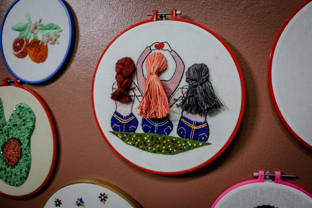 This image shows cross-stich art on a brown wall. The art is inside of a red embroidery hoop and feature three girls sitting on the ground with their backs turned toward our view. | The Ultimate Craft App Guide for Crafters of All Ages | Photo by Ahmed Akacha