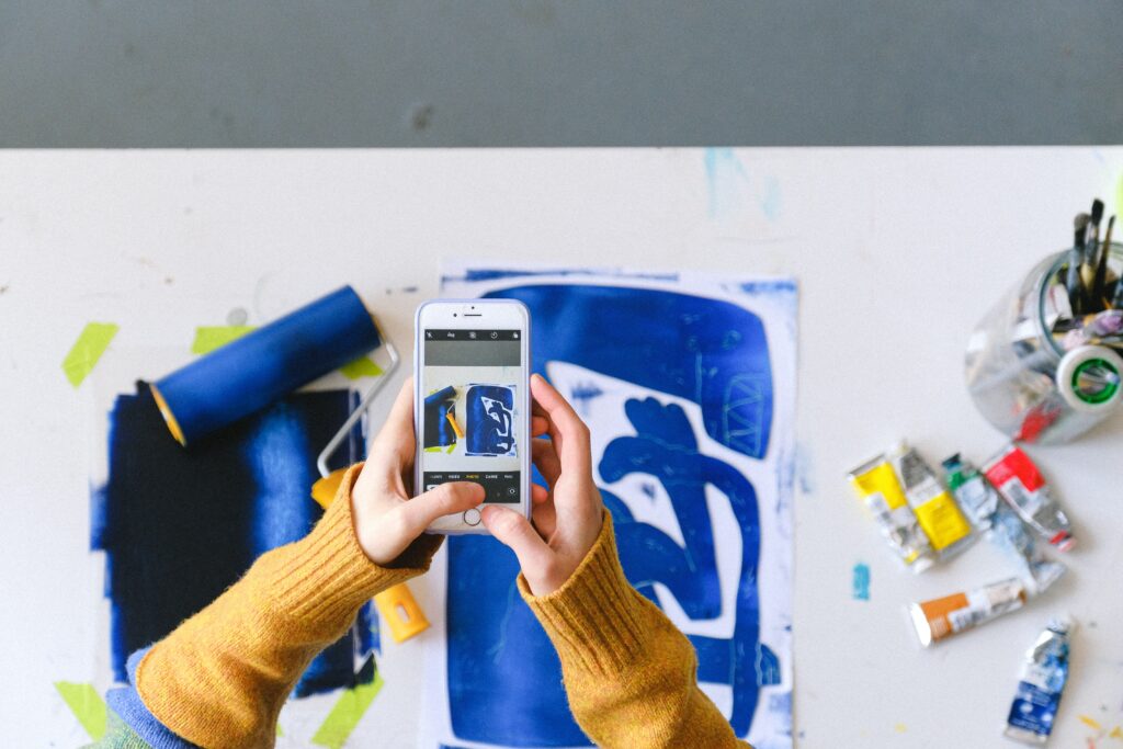 The photo shows a pair of hands taking a picture of blue abstract art laying on a table. | The Ultimate Craft App Guide for Crafters of All Ages | Photo by Anna Shvets