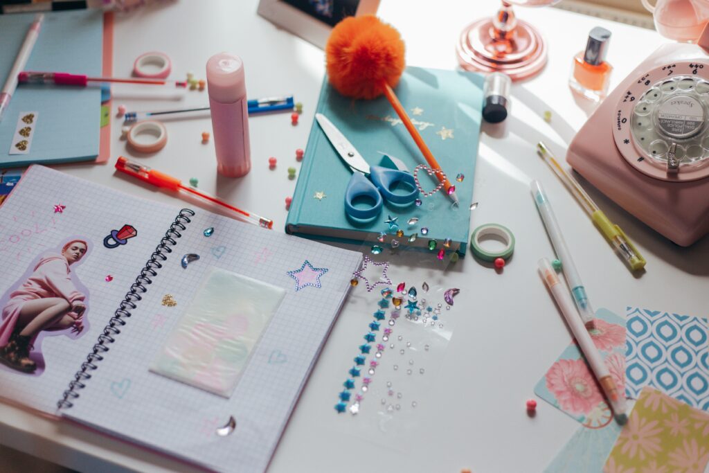 This image shows art supplies scattered across a white desk. The art supplies include a grid notebook, scissors, a pen, sequins, and washi tape | The Ultimate Craft App Guide for Crafters of All Ages | Photo by Cottonbro Studio