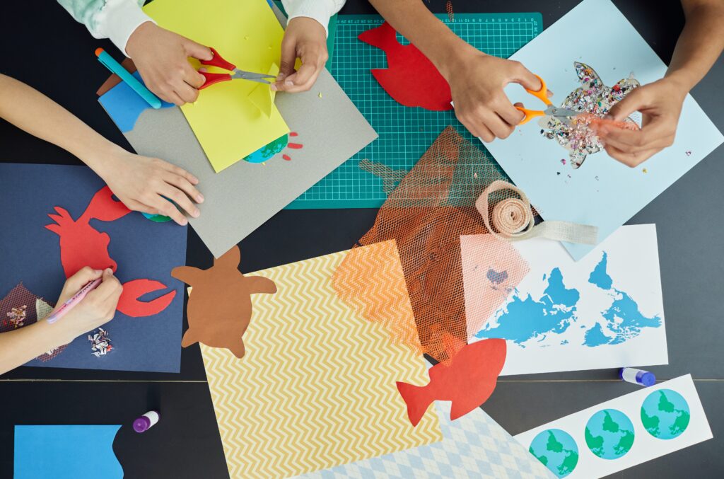 This image is a crop of three pair of kids' hands making crafts at a table covered in paper and paint | The Ultimate Craft App Guide for Crafters of All Ages | Photo by Vanessa Loring