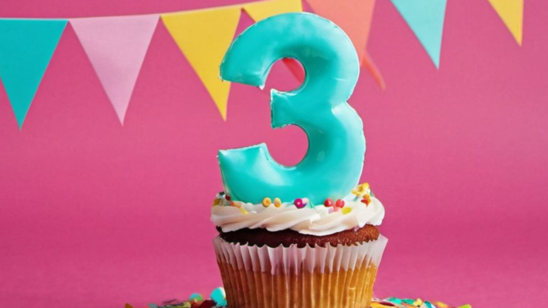 The picture features a turquoise number 3 cupcake with white icing on a pink background, adorned with a multicolor pennant banner and surrounded by vibrant confetti.