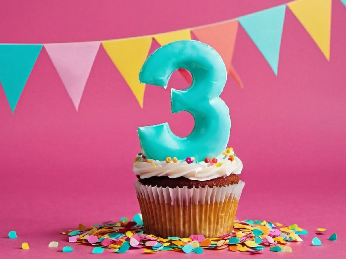 The picture features a turquoise number 3 cupcake with white icing on a pink background, adorned with a multicolor pennant banner and surrounded by vibrant confetti.