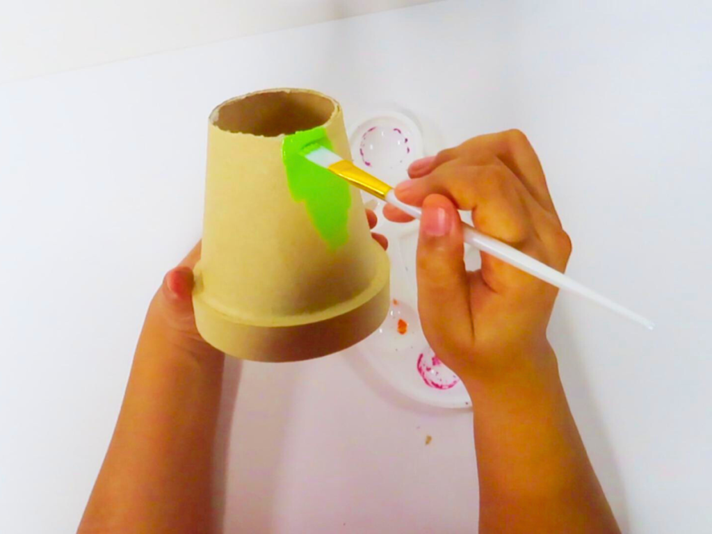 How to Make a Leprechaun Trap for St. Patrick's Day Fun | This image shows a pair of hands painting the paper mache pot with green acrylic craft paint with a white paint brush