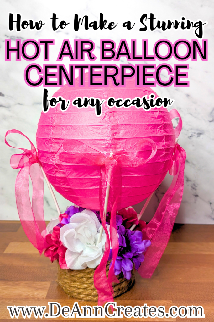 This image is a Pinterest pin entitled "How to Make a Stunning Hot Air Balloon Centerpiece for Any Occasion." It shows a hot air balloon centerpiece with a pink paper lantern as the balloon and it has a basket filled with pink, purple, and artificial flowers. It's sitting on a wooden surface with a white marble background.