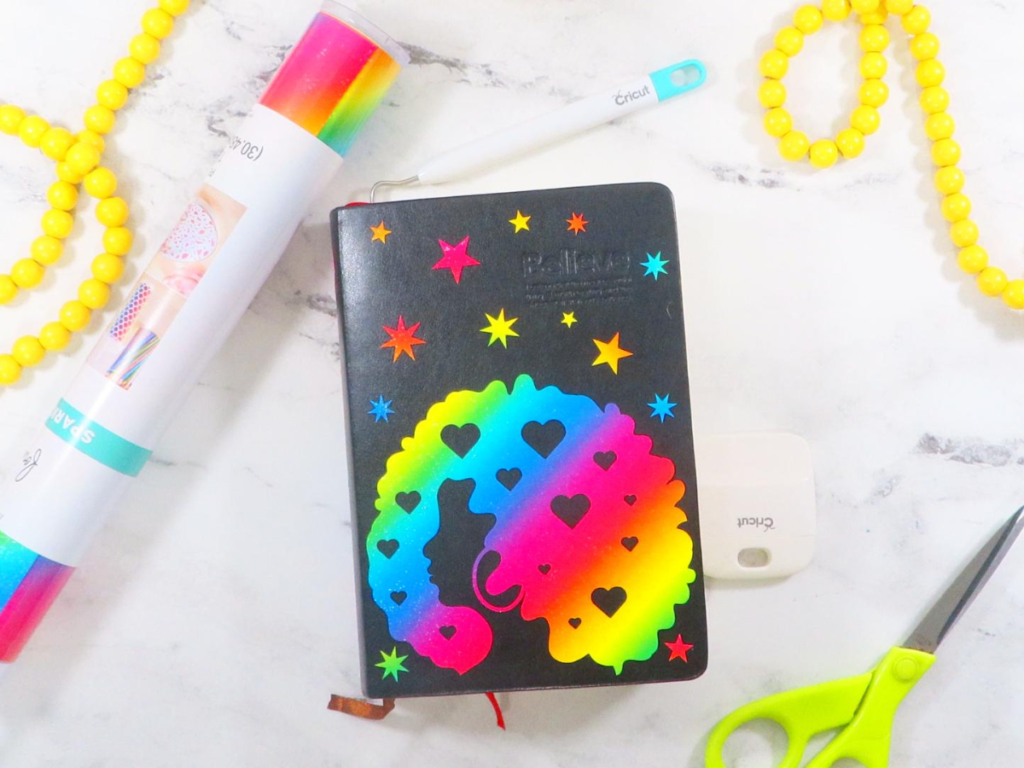 This image shows a bullet journal decorated with rainbow glitter vinyl. The design on the journal features a lady with an afro, stars, and starbursts. The journal is laying on a white marble surface with gray veining. There is also a pair of green scissors, a garland of yellow beads, and a roll of rainbow vinyl laying on the surface. | Personalized Journal Cover Magic with Cricut