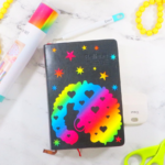 This image shows a bullet journal decorated with rainbow glitter vinyl. The design on the journal features a lady with an afro, stars, and starbursts. The journal is laying on a white marble surface with gray veining. There is also a pair of green scissors, a garland of yellow beads, and a roll of rainbow vinyl laying on the surface. | Personalized Journal Cover Magic with Cricut