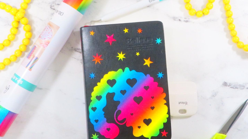 This image shows a bullet journal decorated with rainbow glitter vinyl. The design on the journal features a lady with an afro, stars, and starbursts. The journal is laying on a white marble surface with gray veining. There is also a pair of green scissors, a garland of yellow beads, and a roll of rainbow vinyl laying on the surface. | Personalized Journal Cover Magic with Cricut