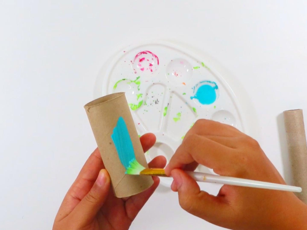 This image shows a hand painting a toilet paper roll turquoise using a white paintbrush | How to Make Toilet Paper Roll Binoculars in a Few Easy Steps