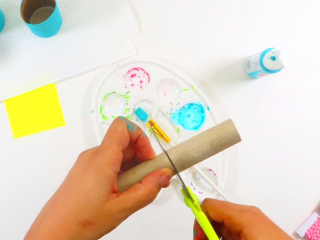 This image shows a hand holding scissors while cutting a skinny cardboard tube | How to Make Toilet Paper Roll Binoculars in a Few Easy Steps