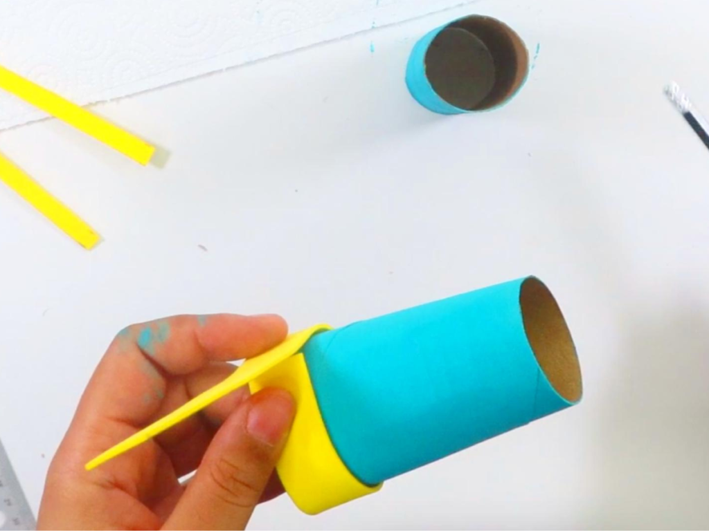 This image shows a hand wrapping the yellow strip of foam around the turquoise painted toilet paper roll | How to Make Toilet Paper Roll Binoculars In a Few Easy Steps