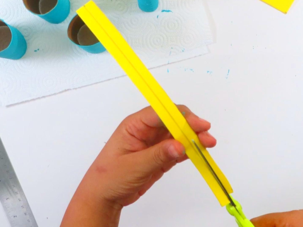 This image shows a hand using scissors to cut two thin strips of yellow foam | How to Make Toilet Paper Roll Binoculars In a Few Easy Steps