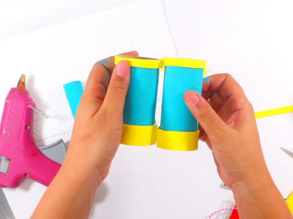 This image shows a pair of hands holding the two painted toilet paper rolls together while letting the hot glue dry | How to Make Toilet Paper Roll Binoculars in a Few Easy Steps