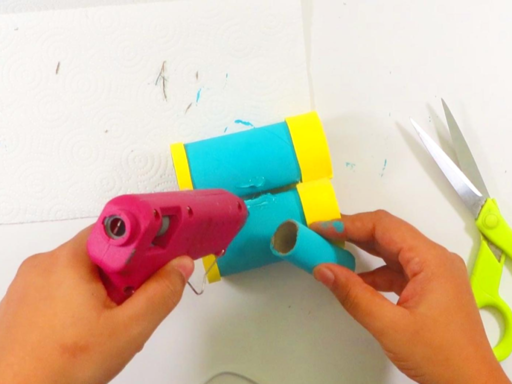 This image shows a pair of hands holding the hot glue gun while gluing the skinny cardboard roll between the two painted toilet paper rolls | How to Make Toilet Paper Roll Binoculars in a Few Easy Steps