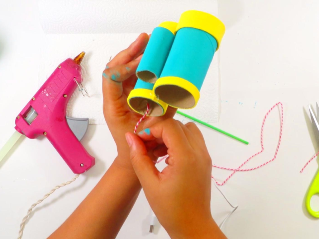 This image shows a pair of hands holding the toilet paper roll binoculars while pressing one end of the decorative twine into the hot glue | How to Make Toilet Paper Roll Binoculars in a Few Easy Steps