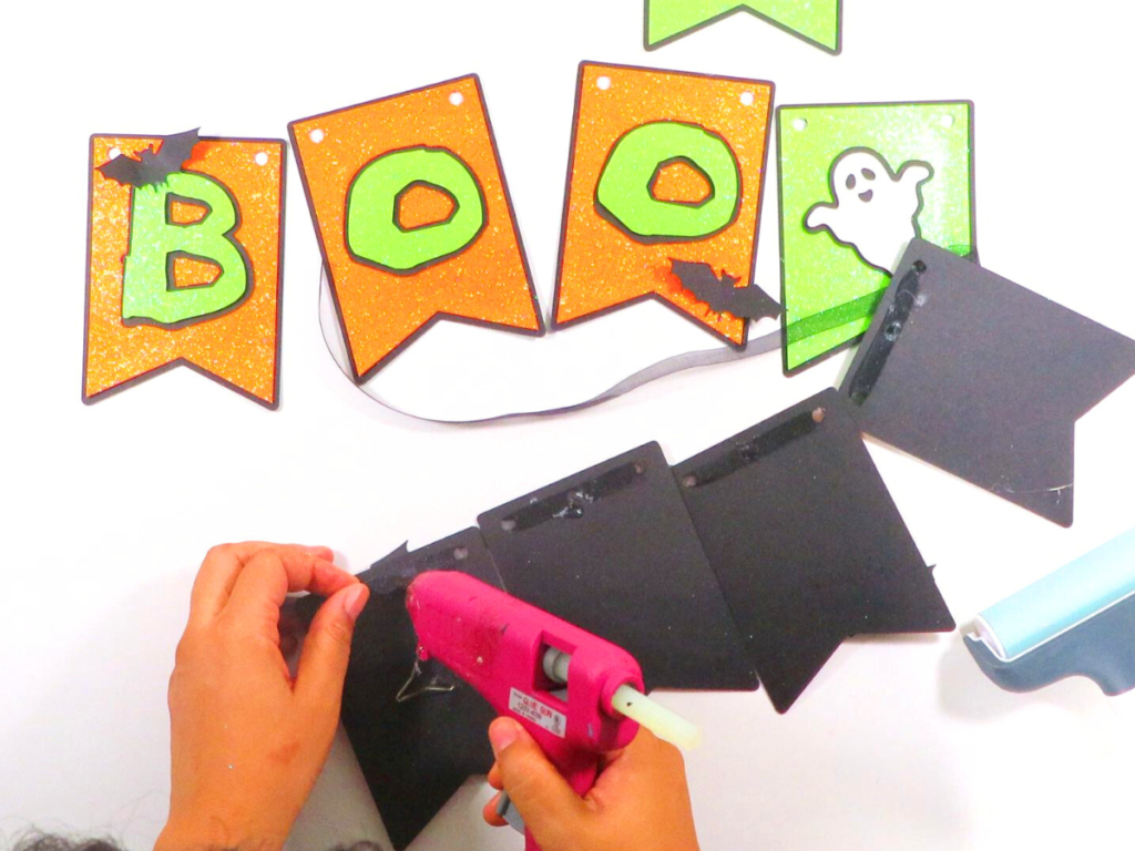 This image shows a pair of hands using a hot glue gun to secure the yarn to the back of each pennant | How to Make a Banner for Halloween That's Cute and Spooky