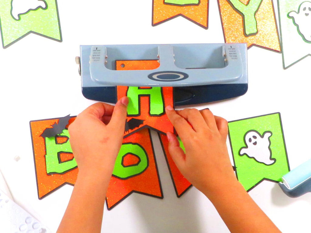 This image shows someone using a hole puncher to punch holes in the top corners of each pennant | How to Make a Banner for Halloween That's Cute and Spooky