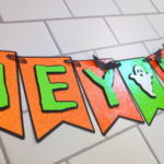 This image shows an orange and green "Hey Boo" Halloween Banner with ghosts and little paper bats. The banner is sitting on a white subway tile background. | How to Make a Banner for Halloween That's Cute and Spooky