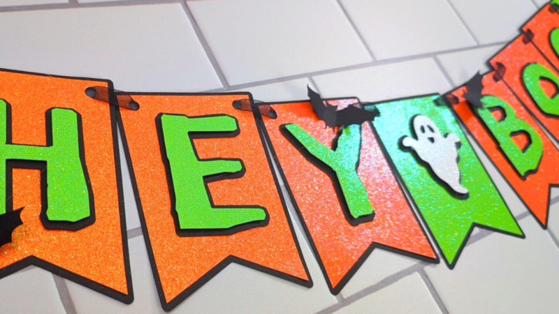 This image shows an orange and green "Hey Boo" Halloween Banner with ghosts and little paper bats. The banner is sitting on a white subway tile background. | How to Make a Banner for Halloween That's Cute and Spooky