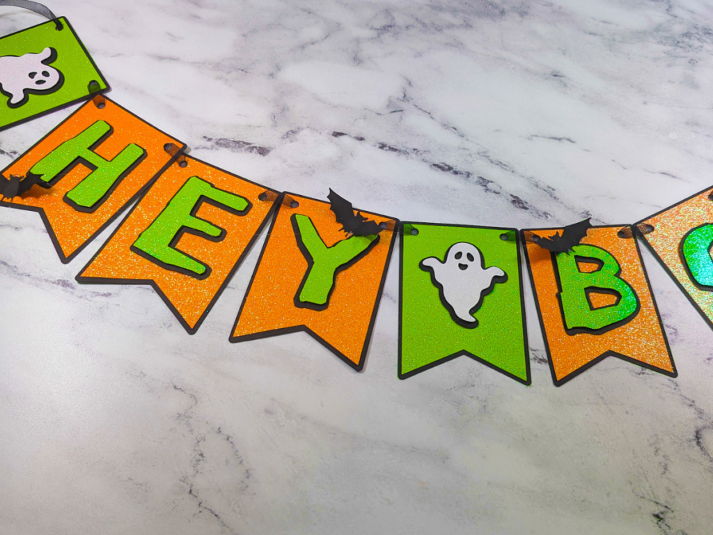 This image shows the finished green and orange "Hey Boo" Halloween banner with ghosts and spiders laying on a white marble background with gray veining. | How to Make a Banner for Halloween That's Cute and Spooky