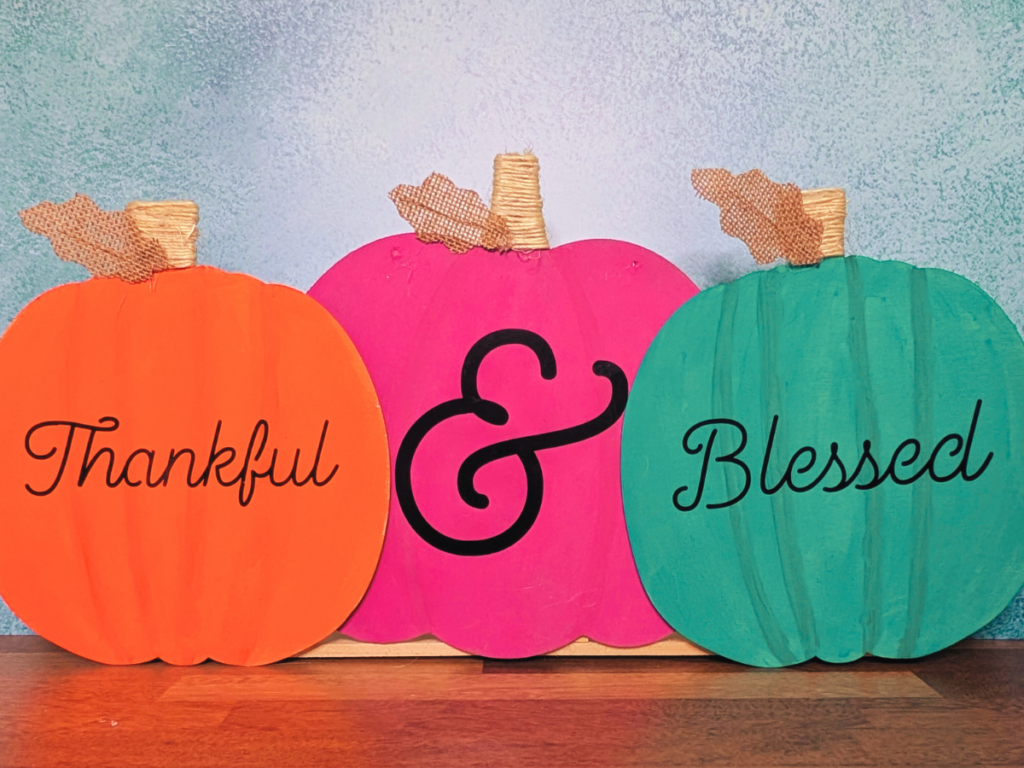 This image shows 3 wooden pumpkins painted orange, pink, and turquoise that have "Thankful & Blessed" written on them with permanent vinyl